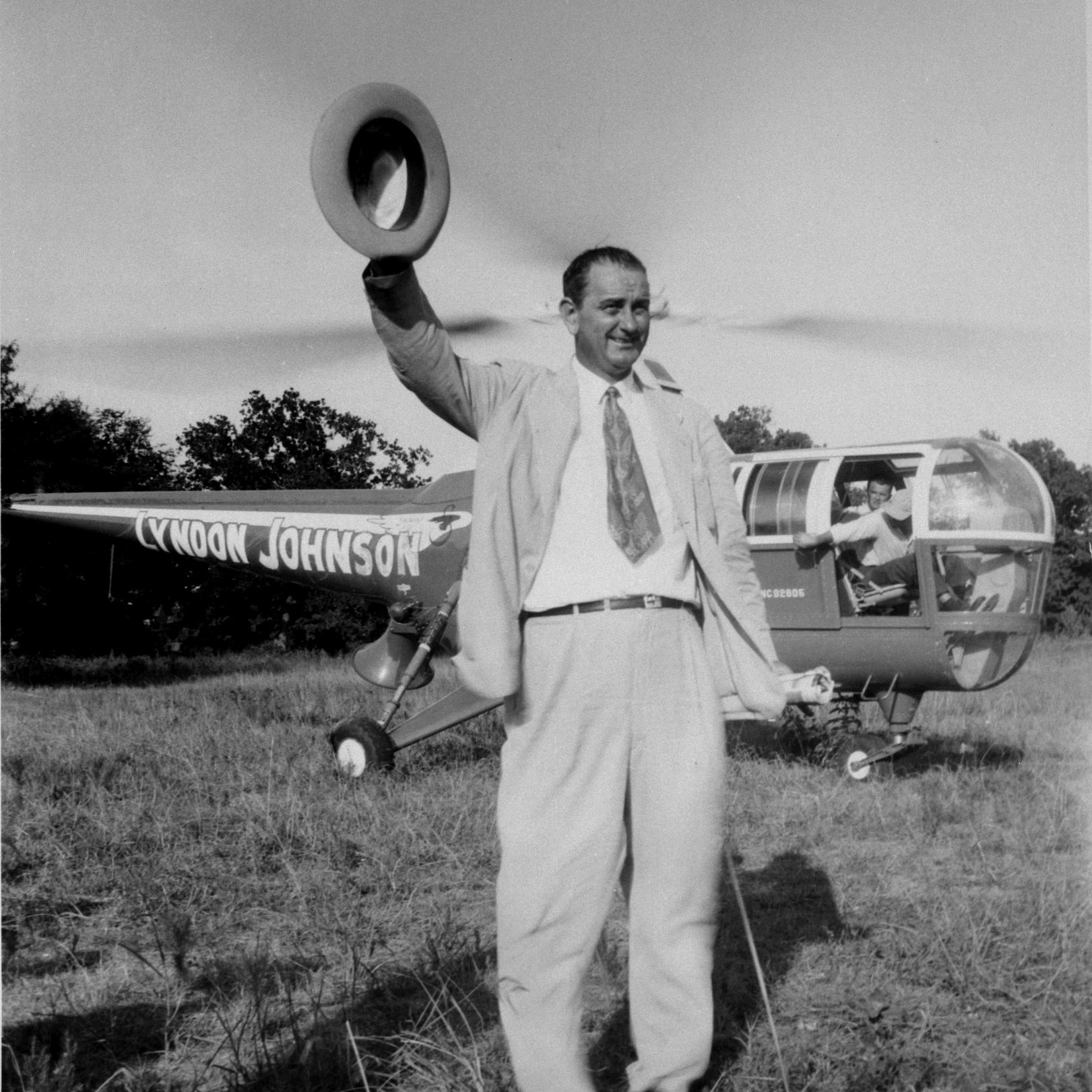 LBJ stands in front of a helicopter with his name on it and waves his hat.